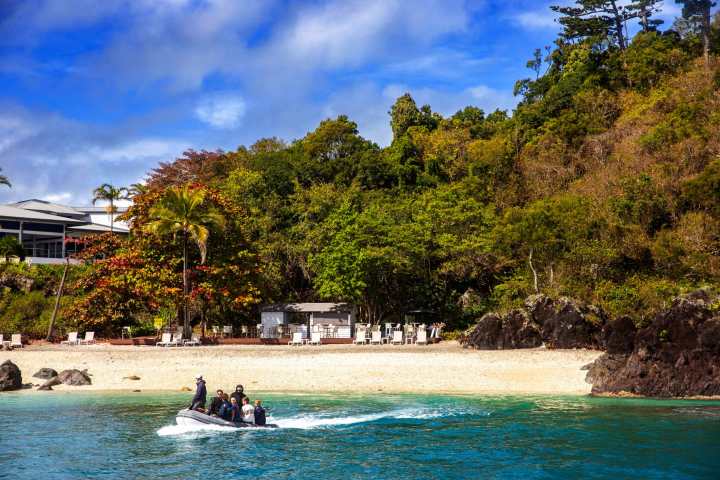 People on a small boat approaching a tropical beach with trees and a building.