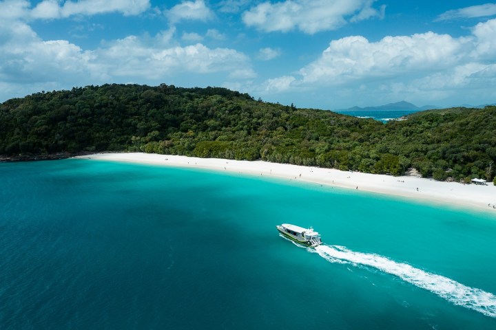 Boat sailing on turquoise water near a sandy beach with lush green forested hills.